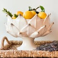 A white ceramic pedestal bowl with a lotus-like petal design, filled with yellow citrus fruits and green leaves, set on a woven wicker tray with dried lavender.