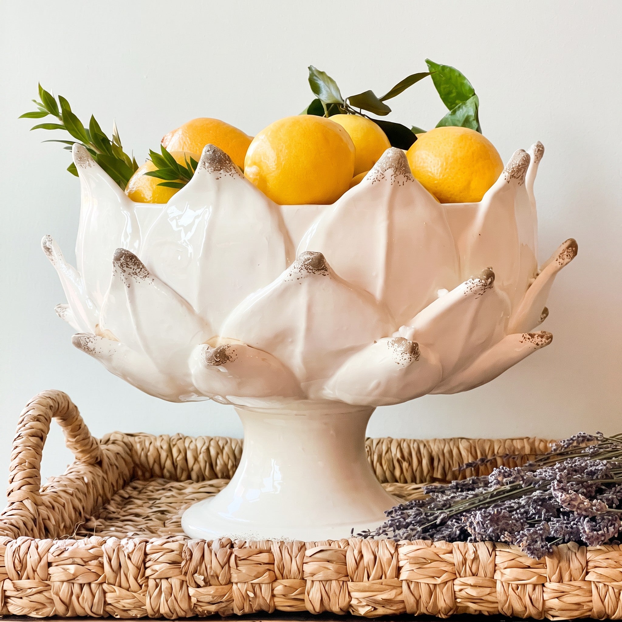 A white ceramic pedestal bowl with a lotus-like petal design, filled with yellow citrus fruits and green leaves, set on a woven wicker tray with dried lavender.